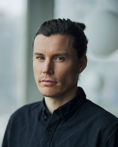 En person med långt brunt hår uppsatt, iklädd en mörk skjorta med knappar, står inomhus framför ett fönster och tittar mot kameran med neutral min.