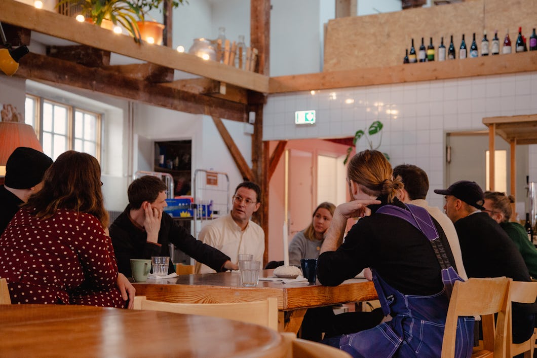 En grupp personer sitter runt ett bord inomhus och pratar. I bakgrunden syns hyllor och flaskor.