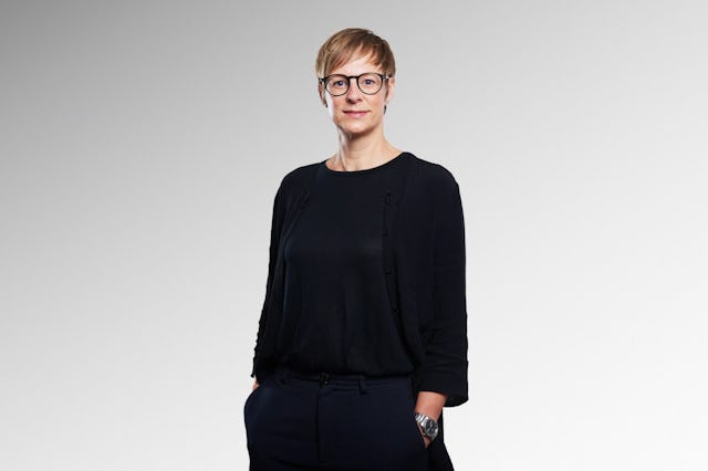Person med kort hår och glasögon står mot en ljus bakgrund, klädd i svart topp, kofta och byxor, med händerna i fickorna.