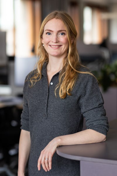 En kvinna med långt blont hår och grå skjorta står inomhus vid en disk och ler mot kameran.