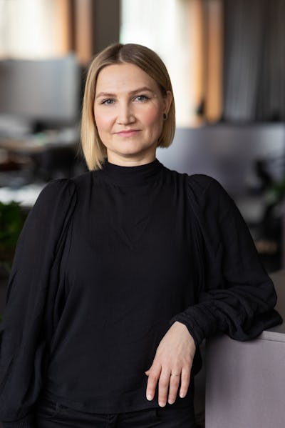 En kvinna med blont hår och svart topp står inomhus med ena armen vilande på en ljusyta och tittar mot kameran.