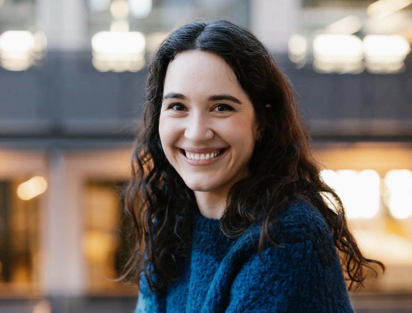 En kvinna med långt lockigt mörkt hår och en blå tröja ler mot kameran. Bakgrunden inomhus är suddig.