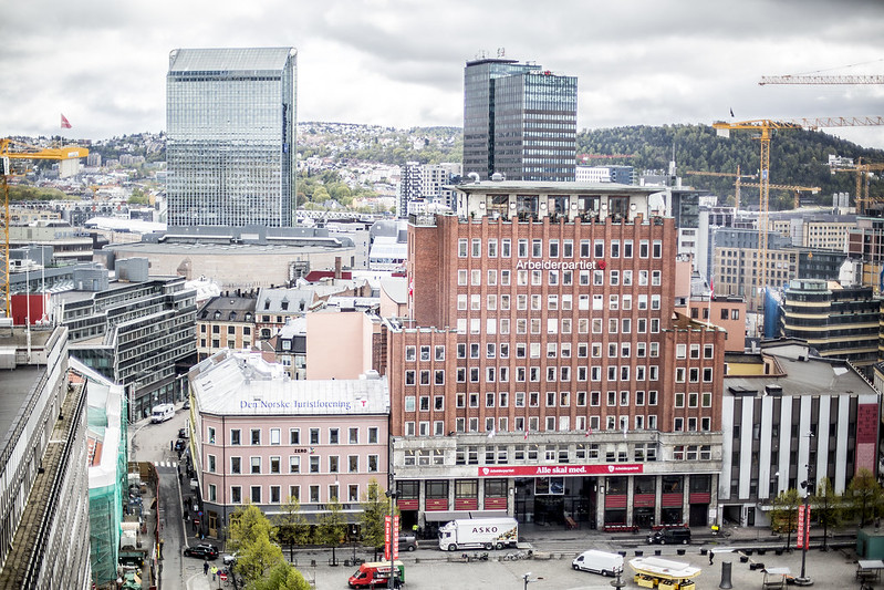 En stadsbild med byggnader från olika tider samt lyftkranar i bakgrunden. Himlen är täckt av moln. Torget nedan används av fordon och fotgängare.