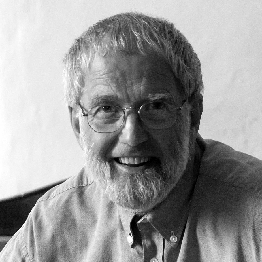 En äldre man med glasögon och skägg ler. Han har en skjorta med knappar och har fotograferats i svartvitt framför en enkel bakgrund.