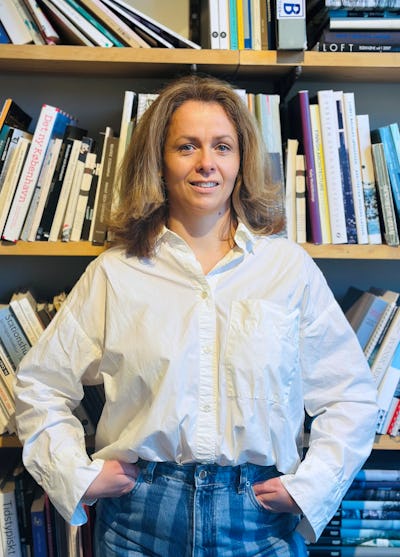 En person med en vit skjorta och blå jeans står med händerna i fickorna framför bokhyllor fyllda med böcker.