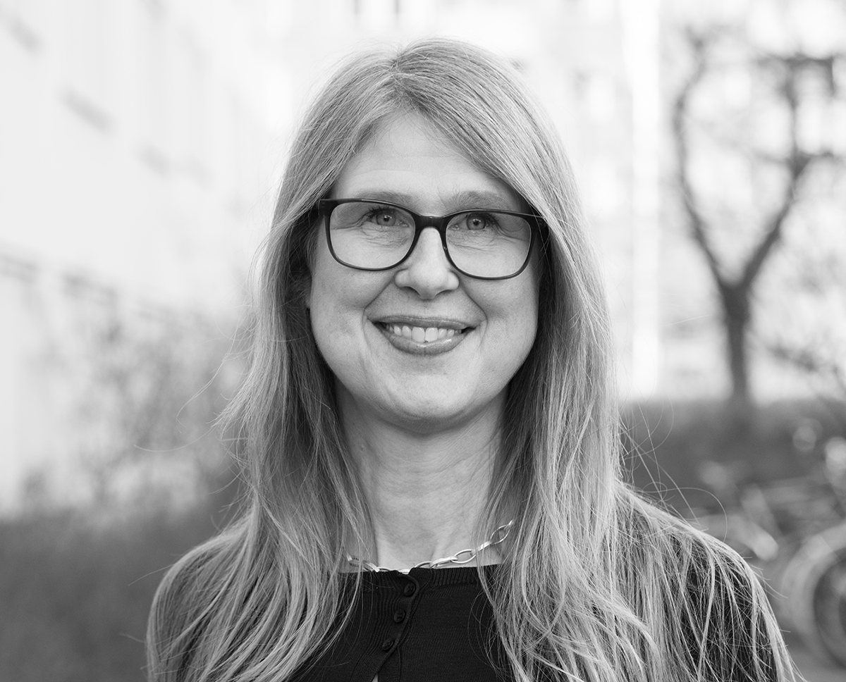 En kvinna med långt hår och glasögon står utomhus och ler mot kameran. I bakgrunden syns byggnader och suddiga träd.