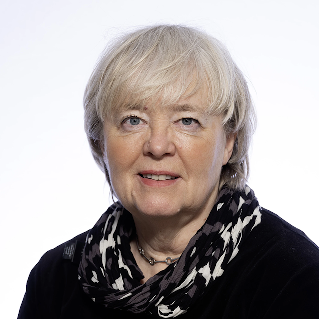 En kvinna med kort, ljust grått hår har på sig en svart topp och en mönstrad scarf. Hon står mot en ljus bakgrund.