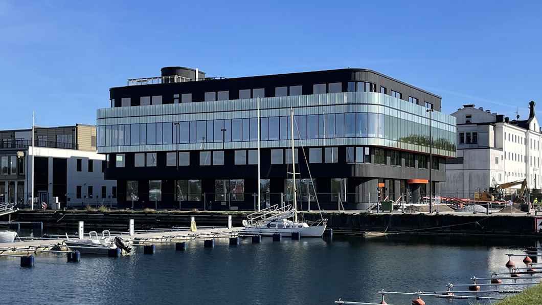 Ett hus av glas och svart färg står vid en marina med tomma bryggor och några båtar under en molnfri himmel.