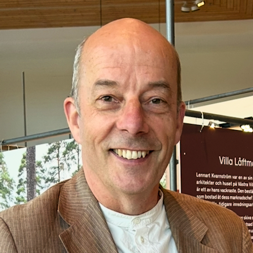 En man i medelåldern med kavaj och vit skjorta står inomhus nära informationspaneler och ler mot kameran.