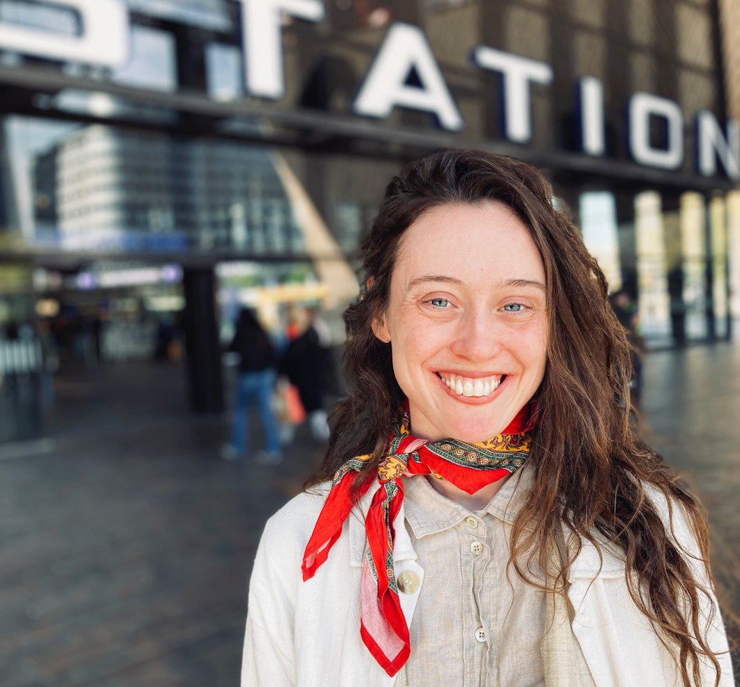 En kvinna med långt brunt hår och en röd halsduk står utanför en ingång med en skylt där det står "STATION" i bakgrunden och ler mot kameran.