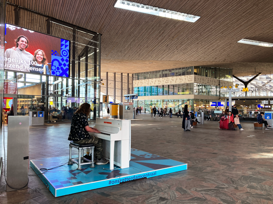 En person spelar piano på en plattform i huvudhallen i en modern tågstation där människor går förbi och en stor digital skärm finns ovanför.