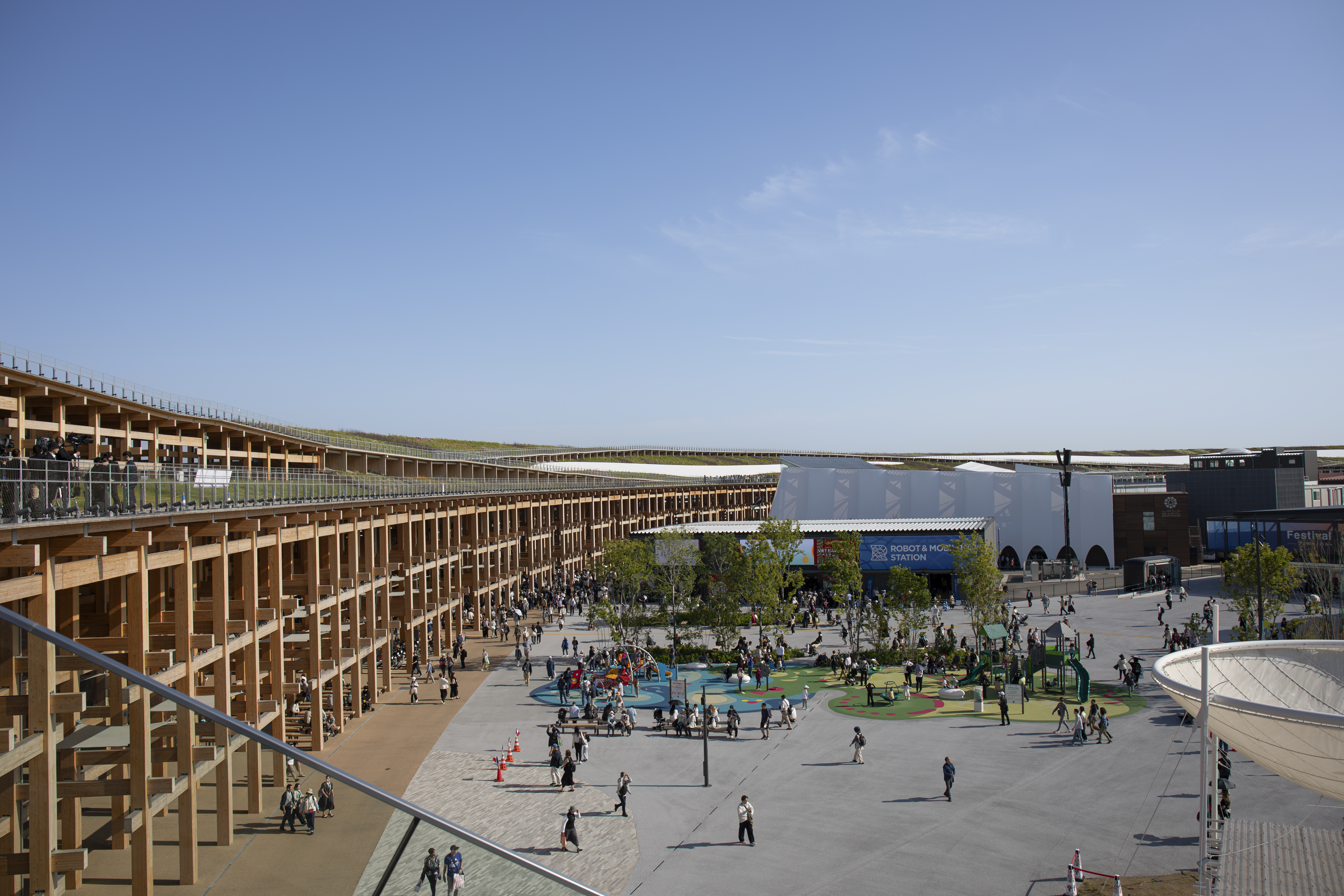 En öppen plats med några människor, omgiven av träbyggnader och grönområden under en molnfri himmel.