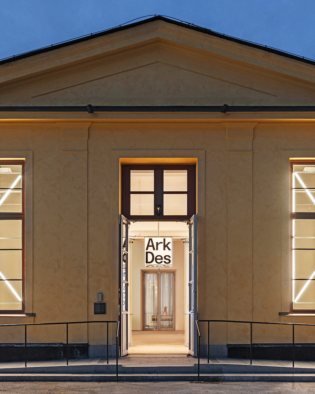 Ingången till ArkDes museums byggnad i skymningen, med belyst interiör och en skylt med namnet "ArkDes" ovanför dörren.