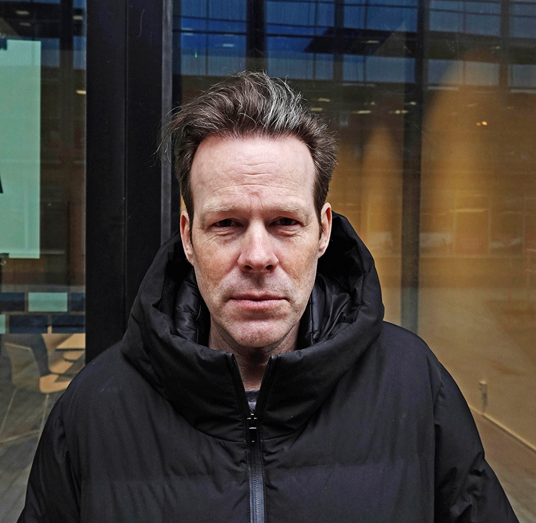 En man med en svart vinterjacka står utanför en byggnad med stora glasfönster och tittar rakt mot kameran.
