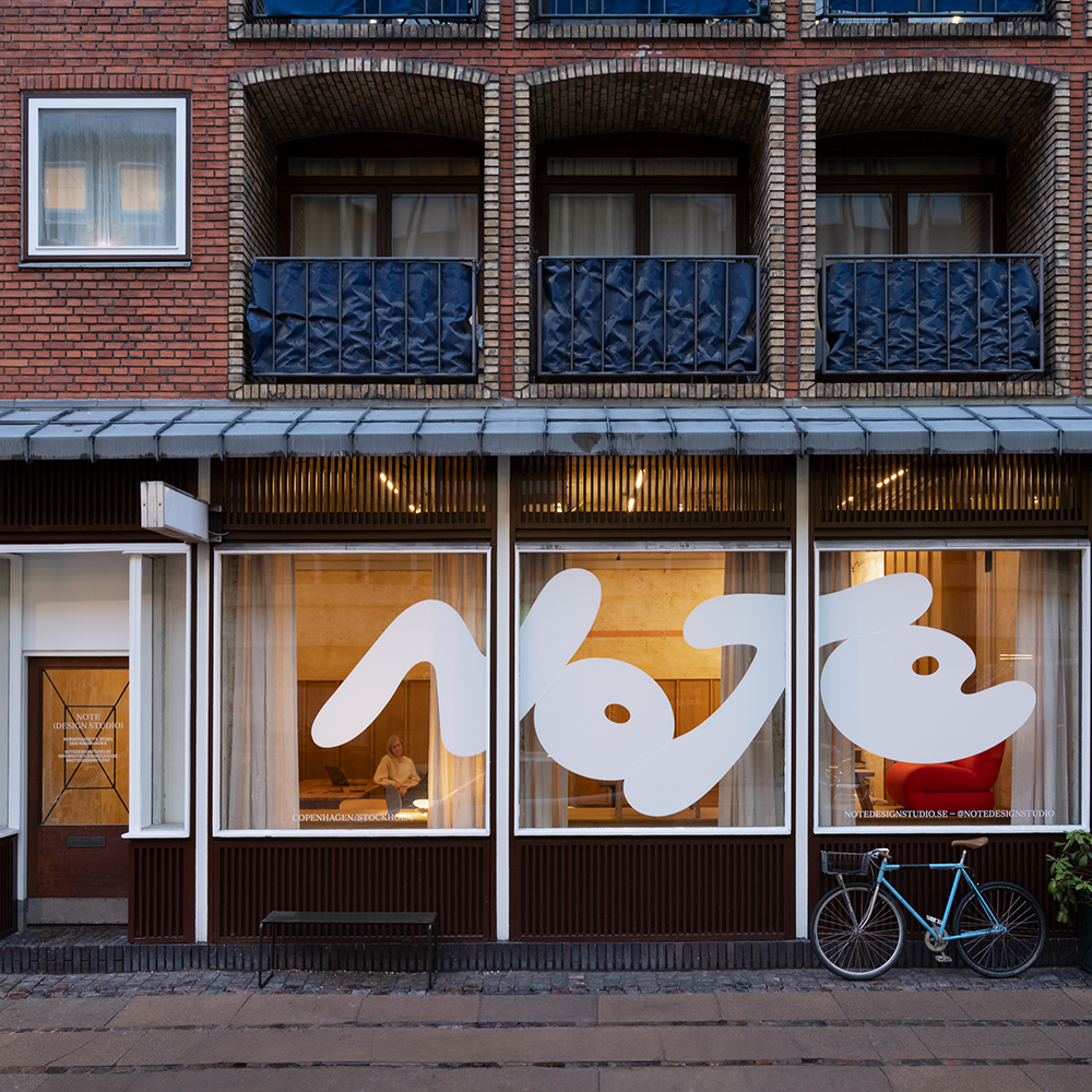 Utsidan av en tegelbyggnad har stora fönster med vita abstrakta mönster. En blå cykel står parkerad på trottoaren framför.