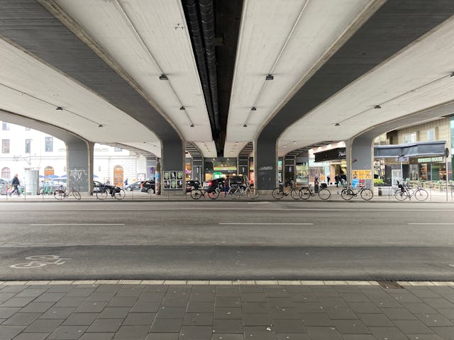 En plats under en bred viadukt med flera pelare. Flera cyklar står parkerade, och några personer promenerar på trottoaren och gatan. I bakgrunden syns butiker och byggnader.