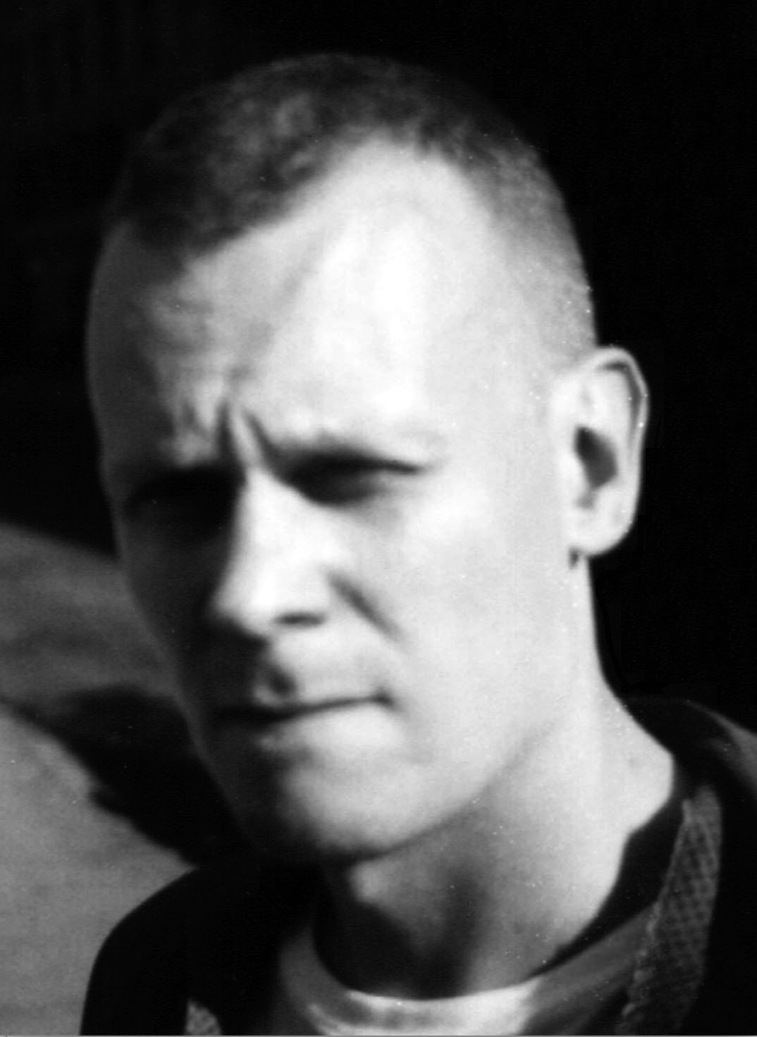 Ett svartvitt foto av en man med kort hår som tittar lite förbi kameran. Han har ett allvarligt ansiktsuttryck och bakgrunden är suddig.