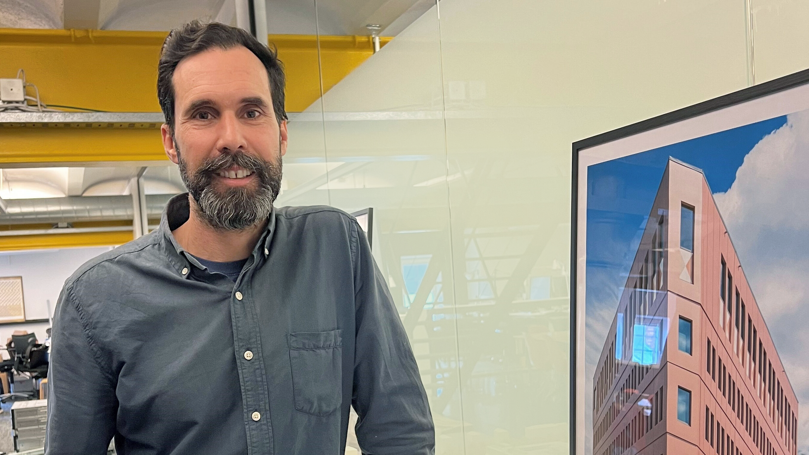 En man med skägg står inomhus bredvid ett inramat fotografi av arkitektur, och i bakgrunden syns och kontorsmiljö.