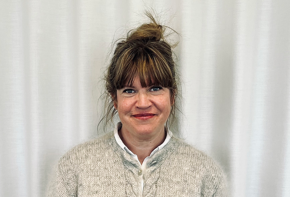 En kvinna med ljusbrunt hår uppsatt i en knut står framför ett vitt draperi. Hon har på sig en stickad tröja och en vit skjorta. Hon tittar mot kameran och ler svagt.