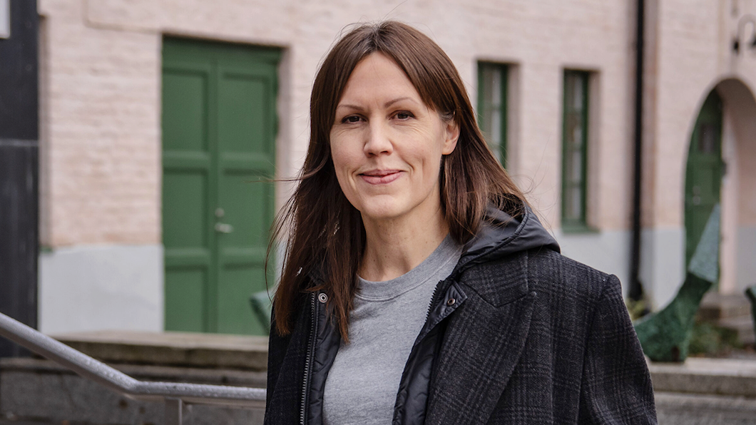 En kvinna med rakt brunt hår, iförd en mörk rock och grå skjorta, står utomhus och ljus byggnad med gröna dörrar.