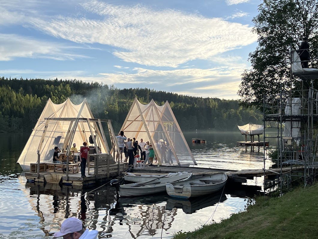 Människor samlas inne i genomskinliga tältsstrukturer som flyter på en sjö. Det finns små båtar och en brygga i närheten, och området är omgivet av träd under en delvis molnig himmel.