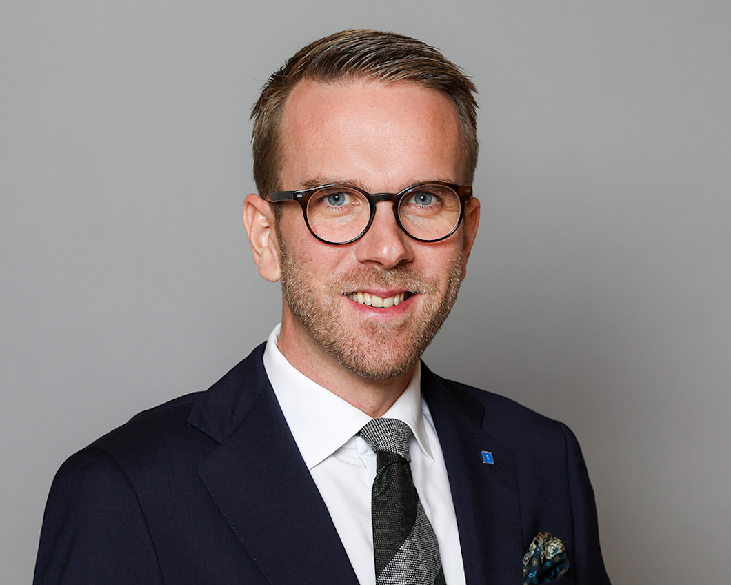 En man med glasögon, kostym, vit skjorta och mönstrad slips står framför en grå bakgrund.
