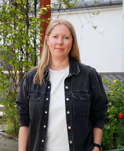 En kvinna med rakt blont hår har på sig en svart jeansskjorta över en vit t-shirt. Hon står utomhus utanför växtlighet och en vit vägg.