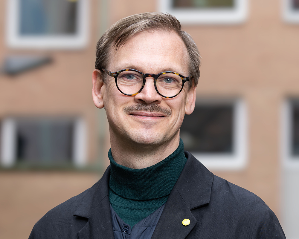 En person med glasögon och mustasch, klädd i en mörk rock och grön polotröja, står utomhus framför en suddig byggnad.