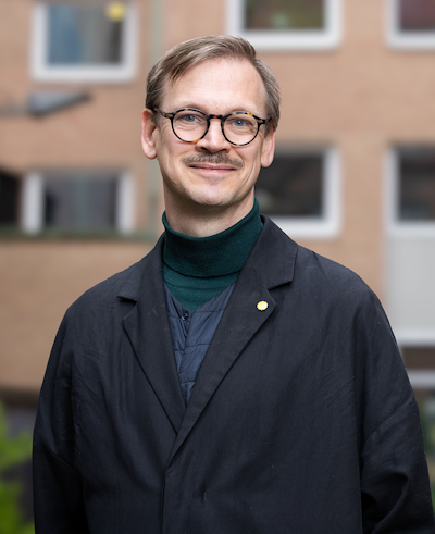 En person med glasögon och mustasch, klädd i en mörk rock och grön polotröja, står utomhus framför en suddig byggnad.