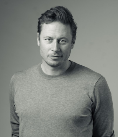En man med ljust hår har på sig en långärmad tröja och tittar mot kameran. Bakgrunden är neutral. Bilden är i svartvitt.