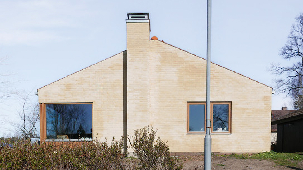 Ett hus byggt av tegel med en skorsten i mitten, två stora fönster och ett slutande tak. Huset ses framifrån med buskar och en gatlykta framför.