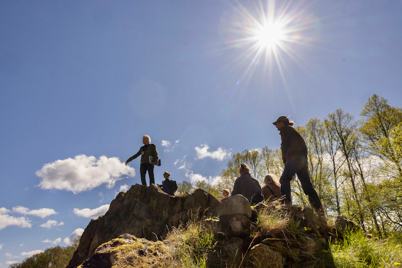En grupp människor står och sitter på en stenig kulle under solen och himlen, med träd i bakgrunden.