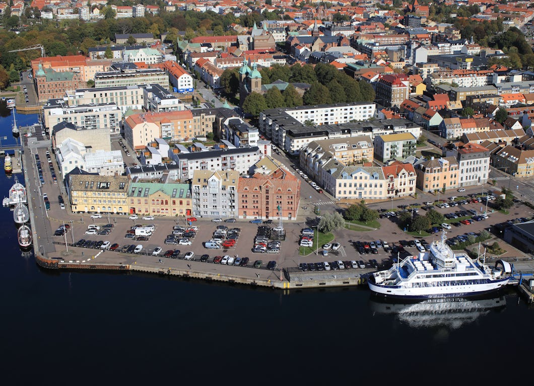 Flygbild över en kuststad med parkerade bilar, en passagerarfärja vid kajen samt bostads- och kommersiella byggnader nära vattnet.