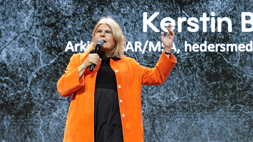 En kvinna i jacka talar på en scen med en mikrofon i handen, framför en skärm som visar namnet "Kerstin B.