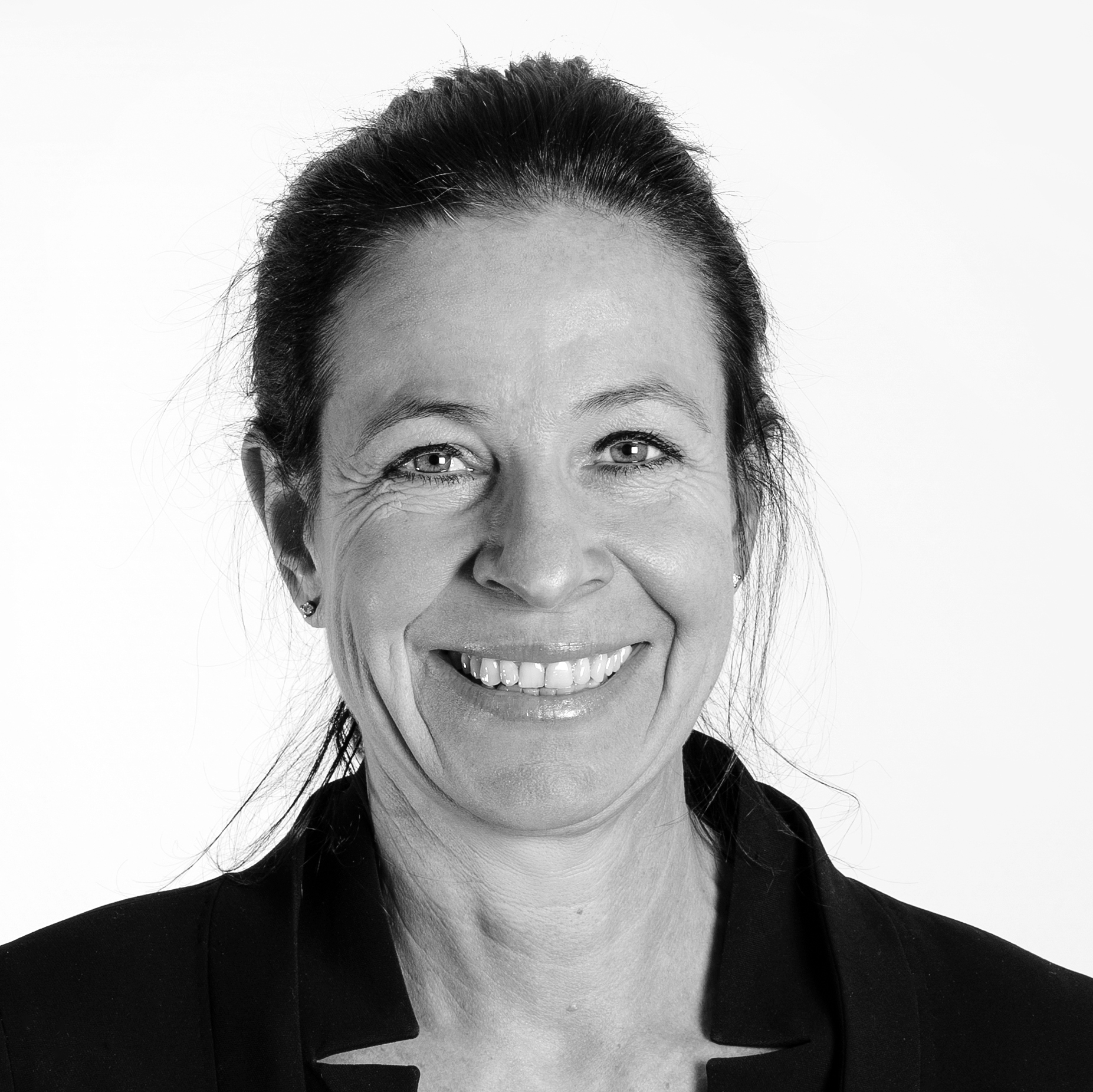 En kvinna med uppsatt hår och en jacka ser in i kameran och ler mot en vit bakgrund.