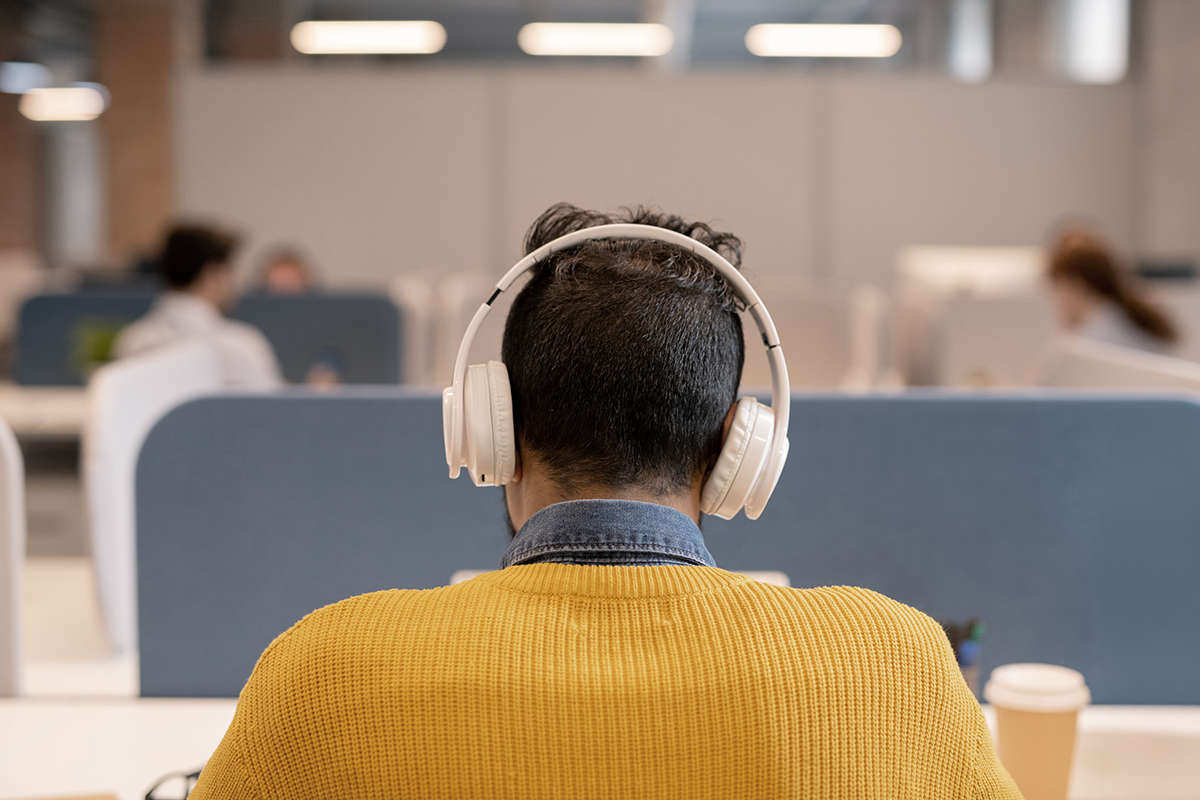 En person med vita hörlurar och gul tröja sitter vid ett skrivbord i ett öppet kontorslandskap, vänd bort från kameran.