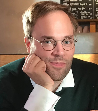 En man med glasögon och ljust brunt hår tittar mot kameran och vill hakan mot handen. Han har en tröja över en vit skjorta med kraft och befinner sig inomhus.
