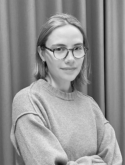 En person med glasögon och axellångt hår har på sig en tröja och står framför ett draperi, vänd mot kameran med ett neutralt ansiktsuttryck.