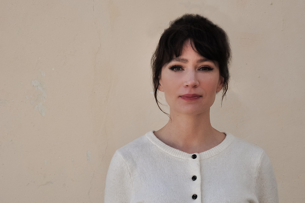 En kvinna med mörkt hår har på sig en vit tröja med knappar och står framför en beige vägg. Hon tittar rakt mot kameran.