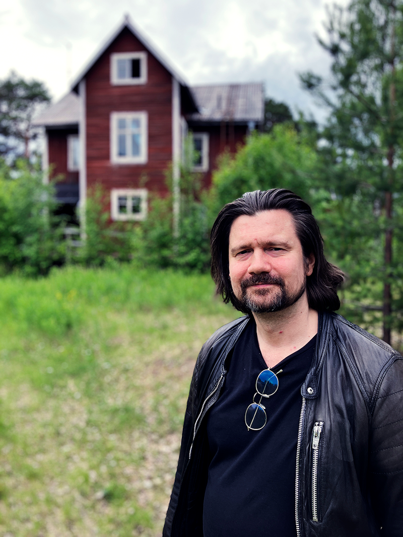 En man med axellångt hår och skägg står utomhus framför ett rött trähus. Han har på sig en svart skjorta, en läderjacka och solglasögon som hänger från skjortan.