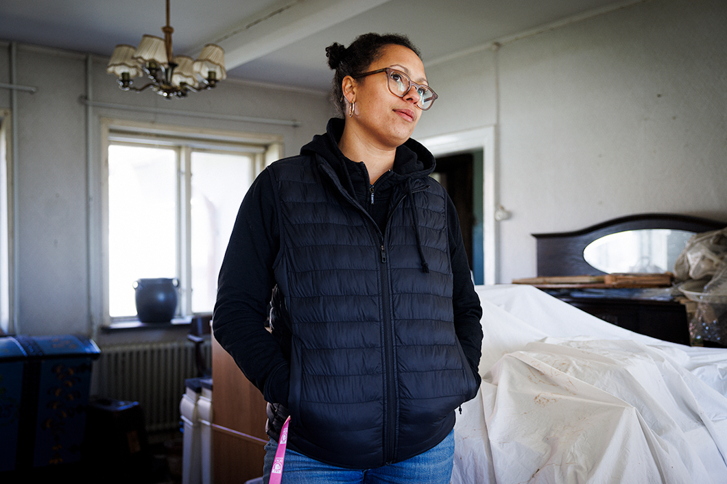 En kvinna med glasögon och svart väst står inomhus med händerna i fickorna, omgiven av möbler och hushållsföremål som är täckta.