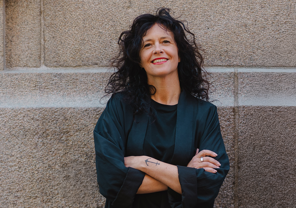 Kvinna med lockigt mörkt hår står mot en stenmur med armarna i kors. Hon bär svarta kläder och har en liten ankartatuering på underarmen.