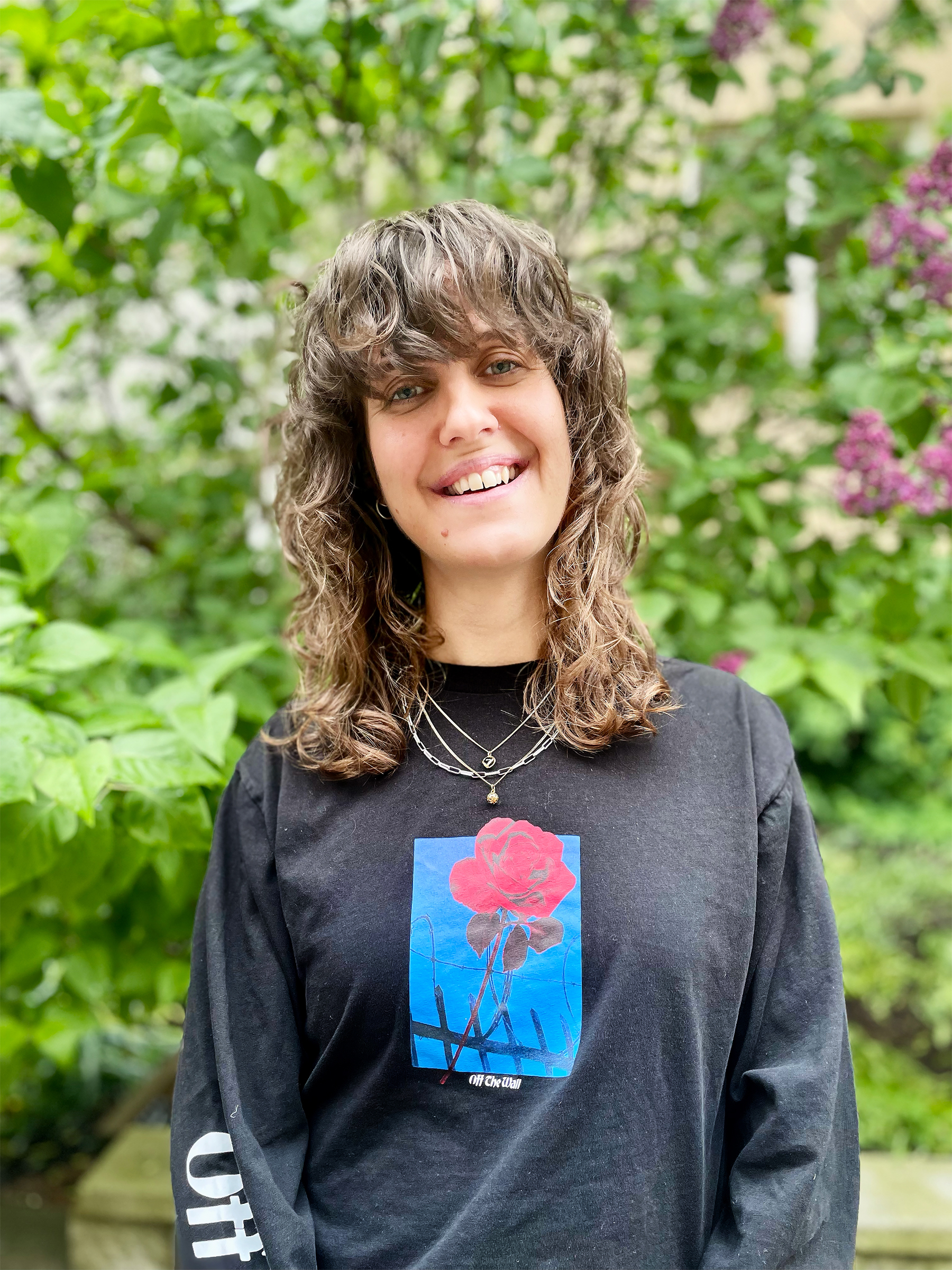 En person med vågigt brunt hår och en svart tröja med ett rött ros-tryck står utomhus framför grönska och lila blommor och ler mot kameran.