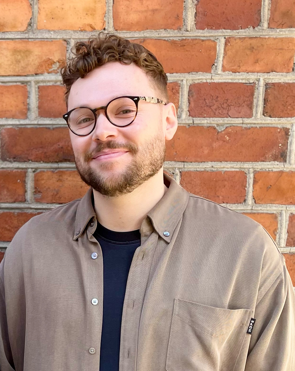 En man med kort lockigt hår, glasögon och skägg står framför en tegelvägg. Han har på sig en brun skjorta över en svart t-shirt.