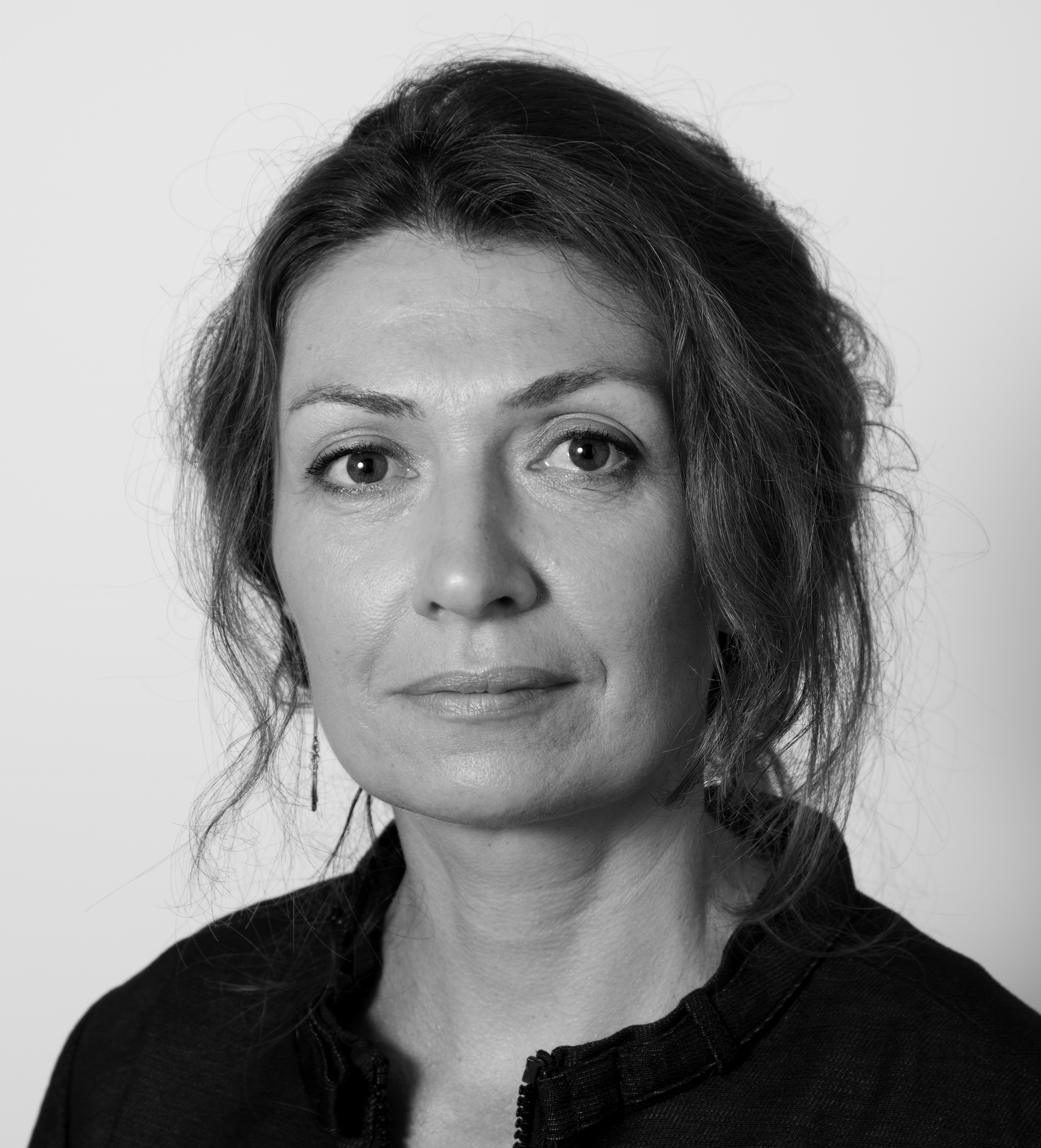 En kvinna med tillbakadraget, vågigt hår och en mörk tröja ser rakt in i kameran med ett neutralt ansiktsuttryck. Bilden är svartvit.