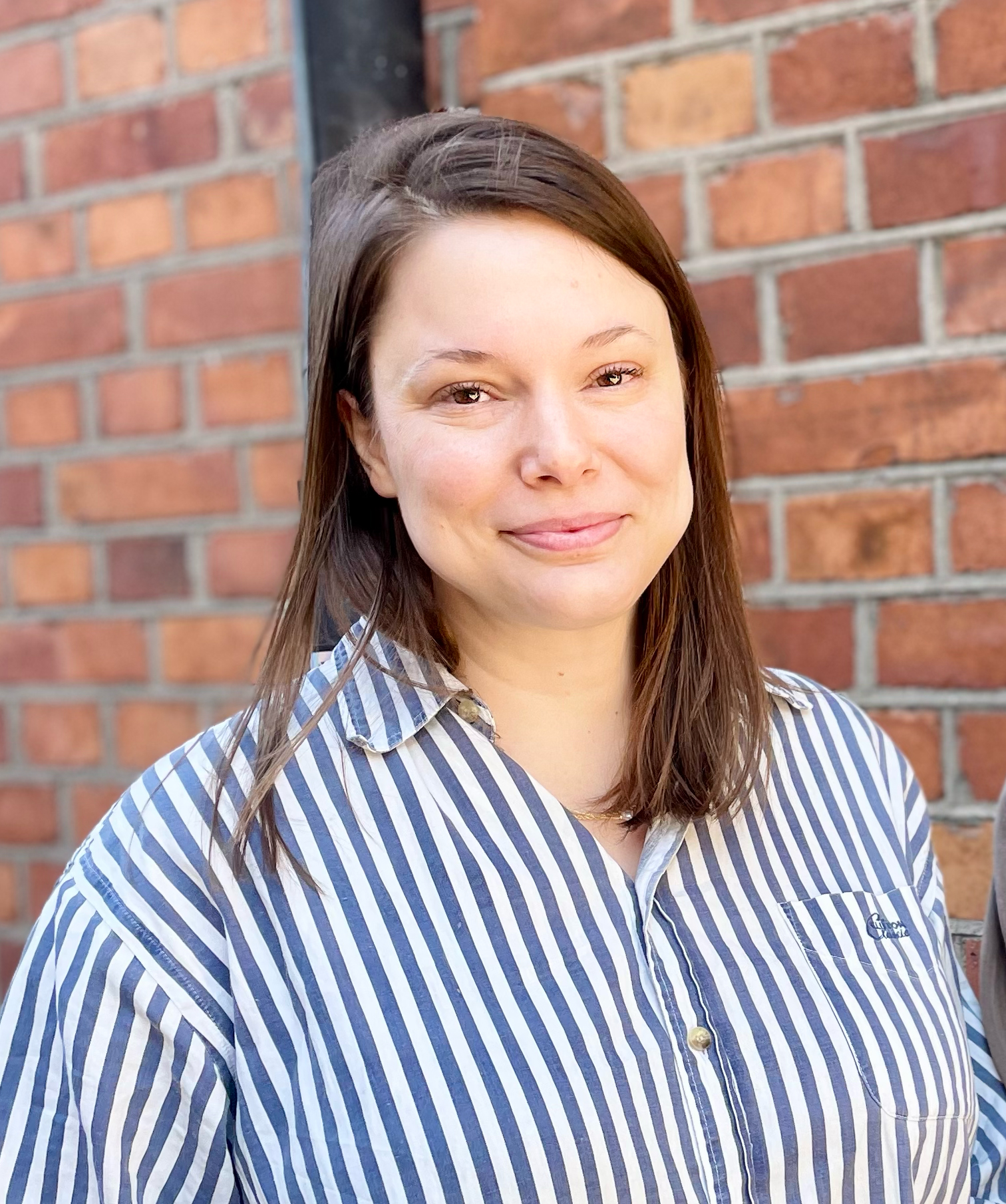 En kvinna med rakt brunt hår och en blåvit randig skjorta står framför en tegelvägg och ler mot kameran.