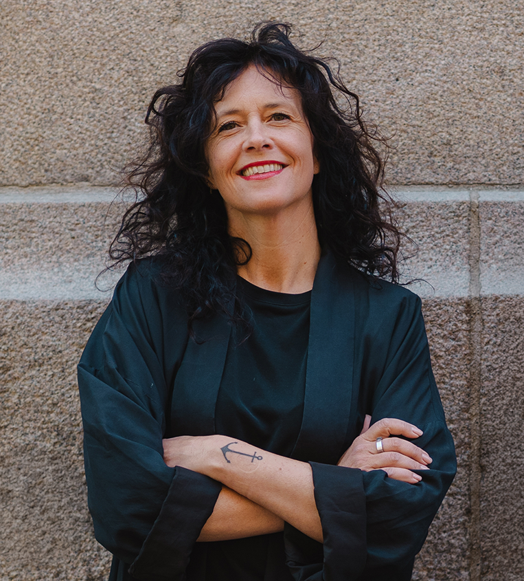 Kvinna med långt, lockigt mörkt hår och en tatuering av ett ankare på underarmen står mot en stenmur. Hon ler, har armarna i kors och bär en svart topp.