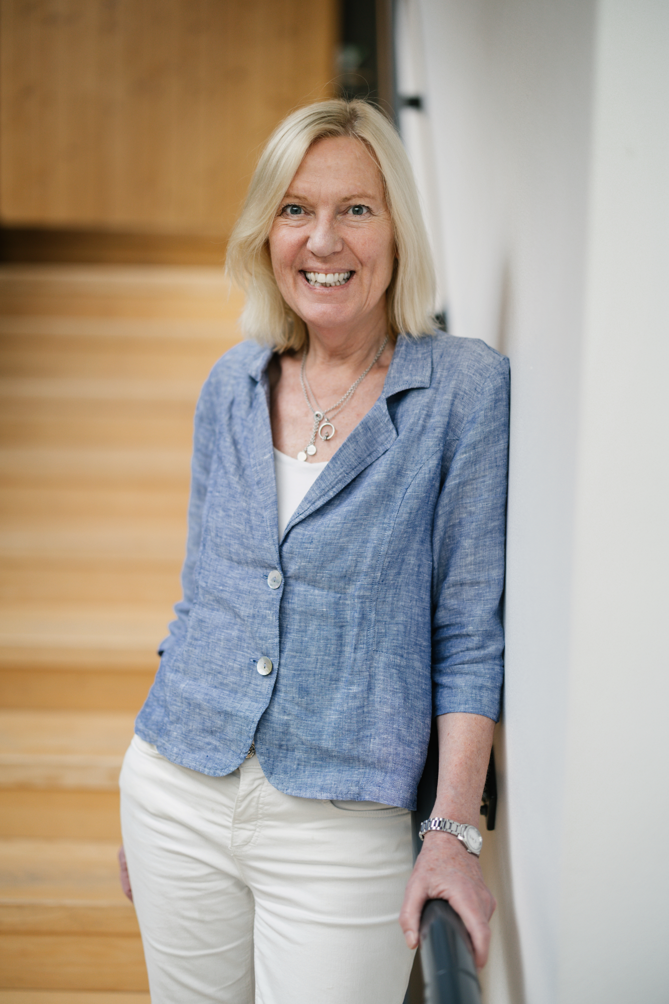 En kvinna med blont hår, iklädd en blå kavaj och vita byxor, står inomhus vid en trappa och ler mot kameran.