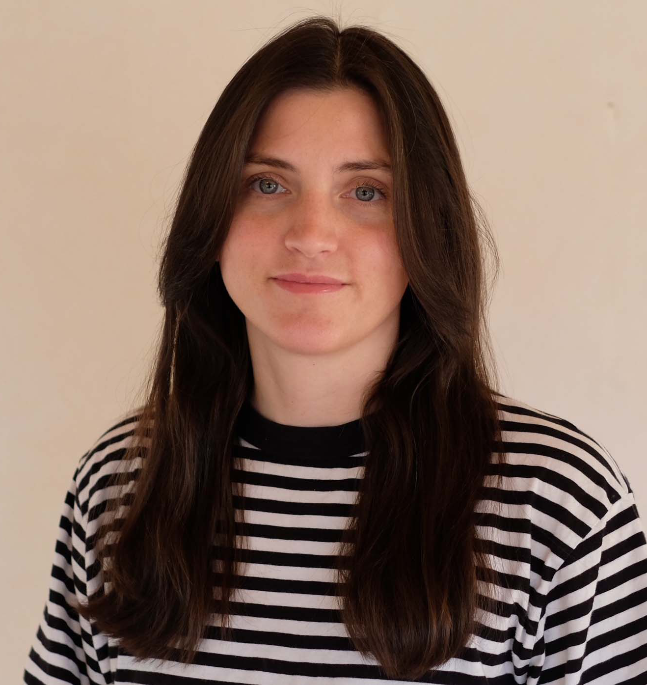 En person med långt brunt hår och blå ögon har på sig en svartvit randig tröja och tittar mot kameran. Personen står framför en beige bakgrund.
