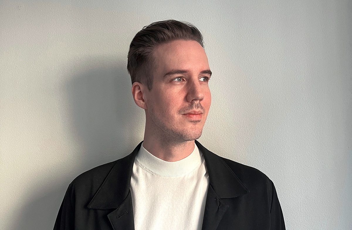 En man med kort hår, klädd i svart jacka och vit polotröja, står mot en ljus vägg och tittar åt vänster.